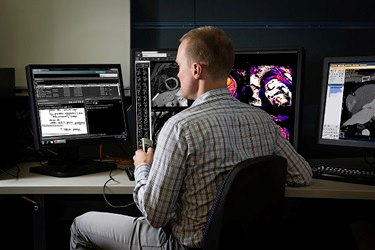 male researcher sitting at desk in front of multiple computure screens looking at imaging.